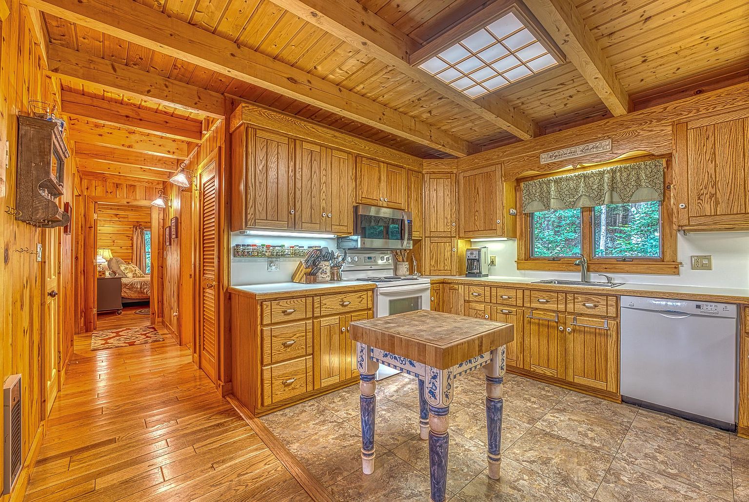 This rustic kitchen features warm wood-paneled walls and ceilings, creating a cozy, cabin-like atmosphere. The space is equipped with light wood cabinetry, a central butcher-block island with decorative painted legs, and a skylight that provides natural illumination. A hallway leads to an adjacent bedroom, emphasizing the open and connected layout of the home.