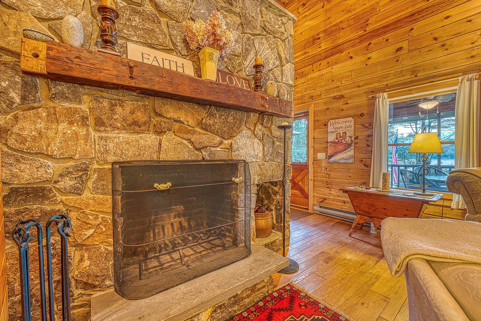 This cozy living room features a prominent stone-clad chimney breast with a rustic timber mantelpiece, serving as the focal point of the space. The room is finished with warm wood-paneled walls, a patterned area rug, and a comfortable seating area near a window that looks out onto a lake. A decorative sign adds a personal touch, creating an inviting and rustic atmosphere typical of a lakeside cabin.