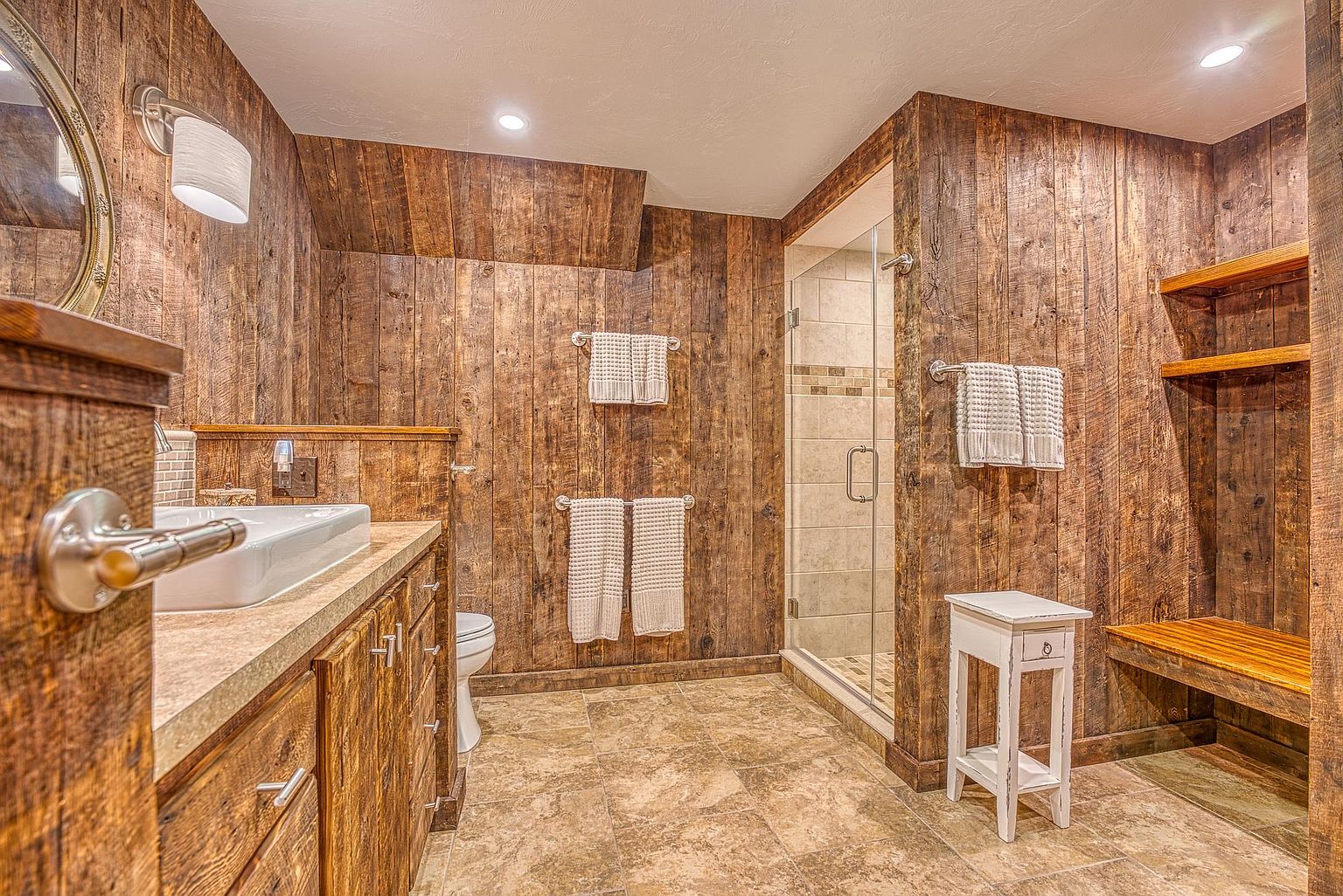 This rustic-style bathroom features extensive wood-paneled walls and a matching vanity with a light-toned stone countertop and a rectangular vessel sink. The space includes a walk-in shower with a glass door, a small white side table, and built-in wooden shelving units. The warm lighting and textured materials create a cozy, cabin-inspired atmosphere.