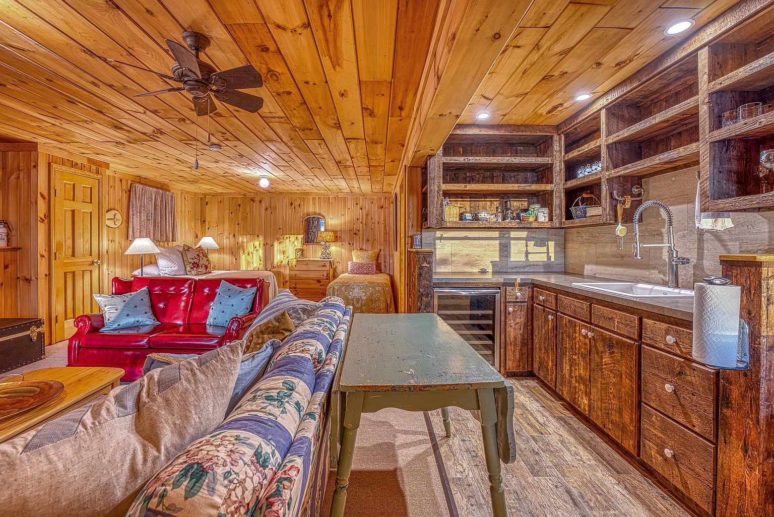 This rustic, open-concept living space features warm wood-paneled walls and ceilings, creating a cozy cabin atmosphere. The room includes a vibrant red leather sofa, a patterned fabric sofa, and a kitchenette area with wooden cabinetry and a stone-tiled backsplash. In the background, a sleeping area with multiple beds is visible, showcasing a versatile layout that blends living and bedroom functions.