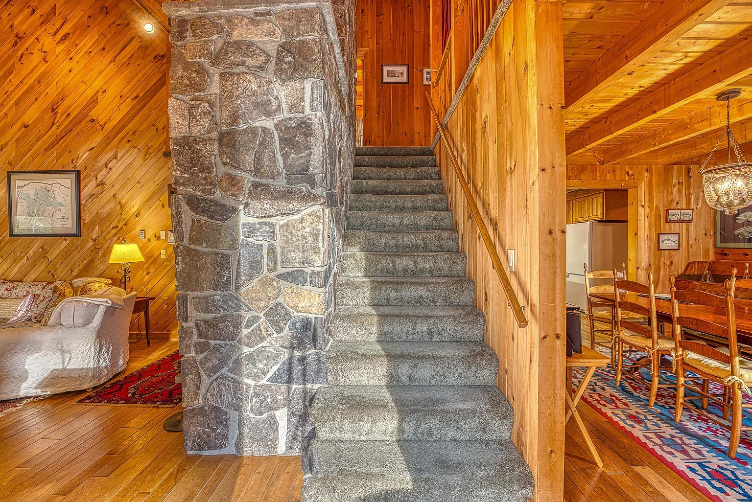This interior view captures a central staircase featuring carpeted steps and a wooden handrail, flanked by a prominent stone-clad chimney breast. To the left, a cozy living area with light wood walls and a patterned rug is visible, while to the right, a dining space with a wooden table and a decorative chandelier opens up. The warm, rustic aesthetic is enhanced by the extensive use of natural wood paneling throughout the home.