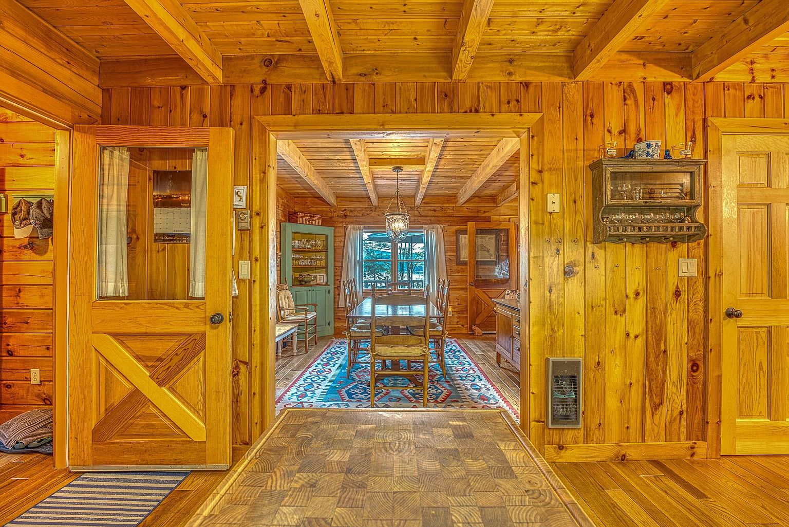 This view from a kitchen island looks through a doorway into a rustic dining room characterized by warm, natural wood walls and exposed ceiling beams. The dining area features a wooden table set for guests, a vibrant patterned area rug, and a decorative hanging light fixture, all framed by windows that look out onto a wooded landscape. The overall aesthetic is cozy and traditional, emphasizing the cabin-like charm of the home's interior architecture.