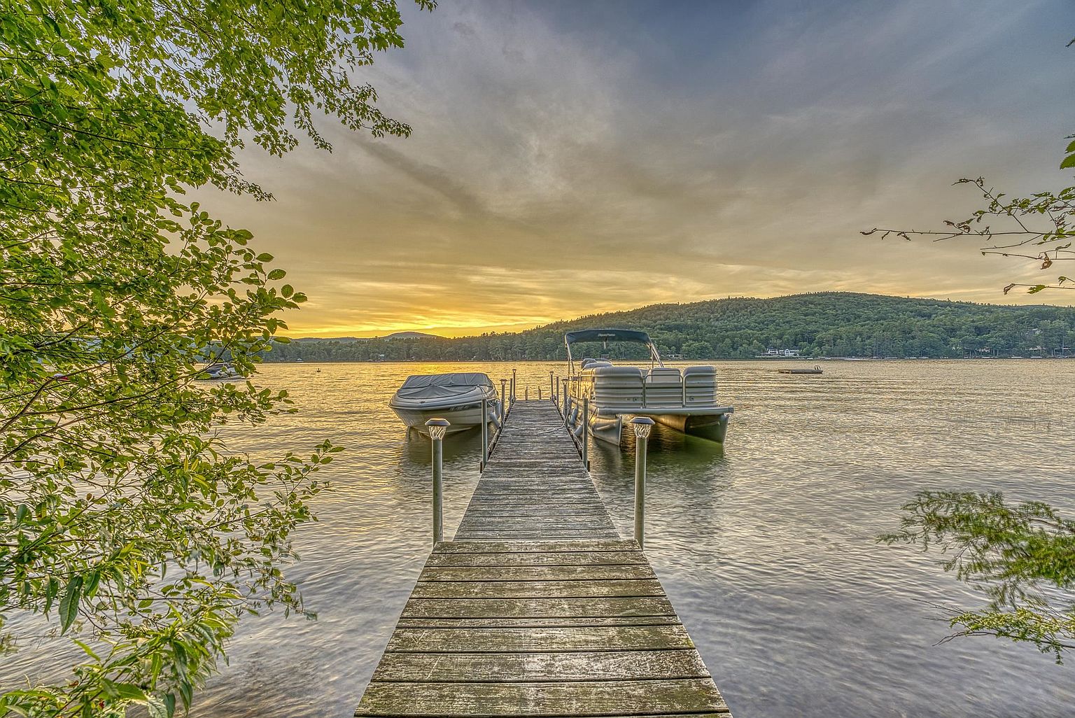This scenic view captures a wooden dock extending into a calm lake at sunset, flanked by two boats moored on either side. The composition features lush green foliage framing the foreground, leading the eye toward the distant, tree-covered hills under a soft, golden-hued sky. The perspective is centered and inviting, highlighting the tranquil waterfront lifestyle and recreational access associated with the property.