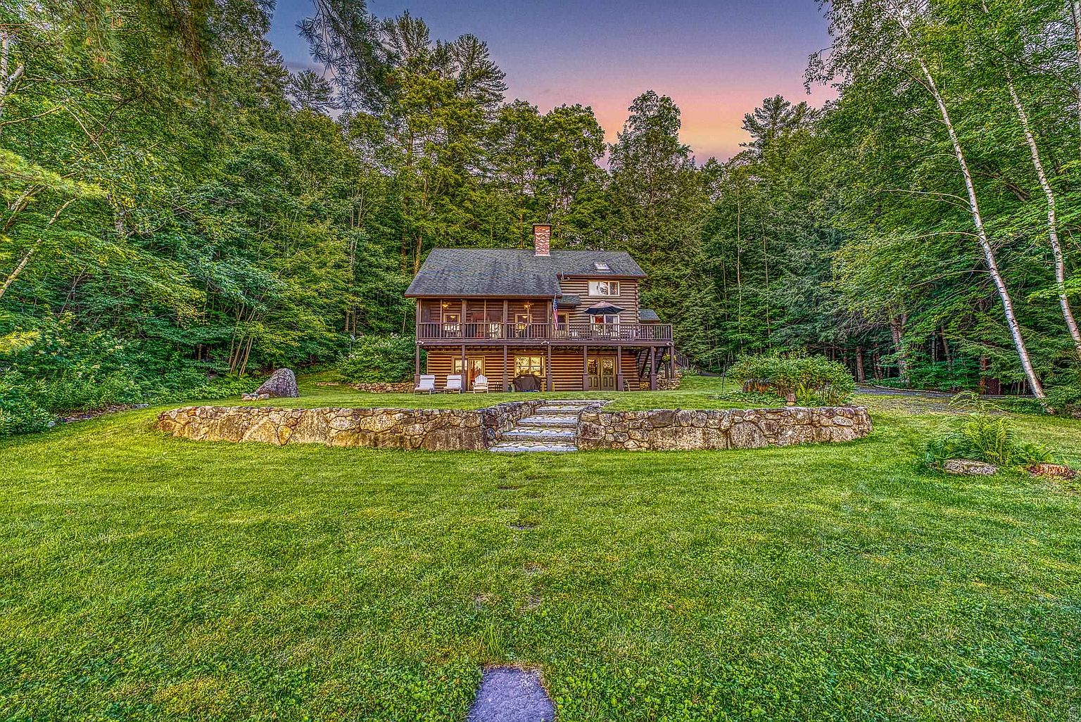 This charming log cabin is nestled within a lush, dense forest, offering a serene and private retreat. The home features a multi-level deck and a stone retaining wall that creates a tiered transition from the expansive, well-manicured lawn to the residence. The warm glow of the setting sun illuminates the sky above the trees, highlighting the rustic architectural character of the structure.