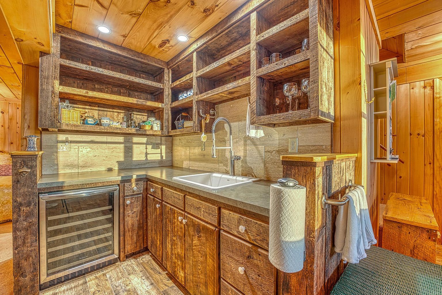 This rustic-style kitchenette features custom reclaimed wood cabinetry and open shelving, creating a warm, cabin-like atmosphere. The space is equipped with a stainless steel wine cooler, a white undermount sink with a gooseneck faucet, and a neutral stone-tiled backsplash. The perspective is a straight-on eye-level shot that highlights the compact, functional layout and the natural wood textures throughout the room.
