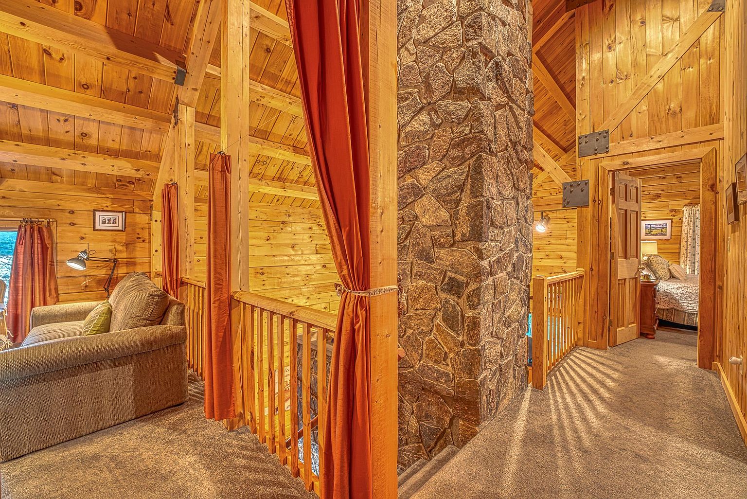 This interior view captures a rustic loft-style hallway featuring warm wood-paneled walls and a prominent stone-clad chimney breast that serves as a central architectural anchor. To the left, a cozy seating area is partially partitioned by orange curtains, while a doorway on the right leads into a bedroom. The space is characterized by its exposed timber framing and a neutral carpet, creating a warm and inviting cabin atmosphere.