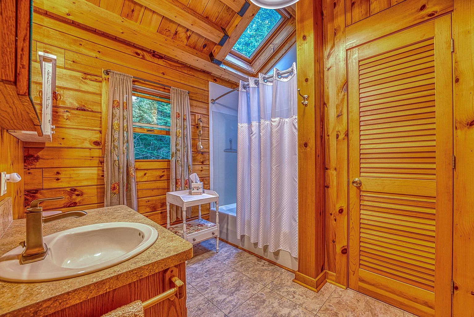 This rustic-style bathroom features warm wood-paneled walls and a matching wood ceiling with a skylight that brings in natural illumination. The space includes a vanity with a stone-look countertop, a shower-tub combination with a white curtain, and a small decorative side table. The room is finished with neutral patterned floor tiles, creating a cozy and inviting atmosphere.