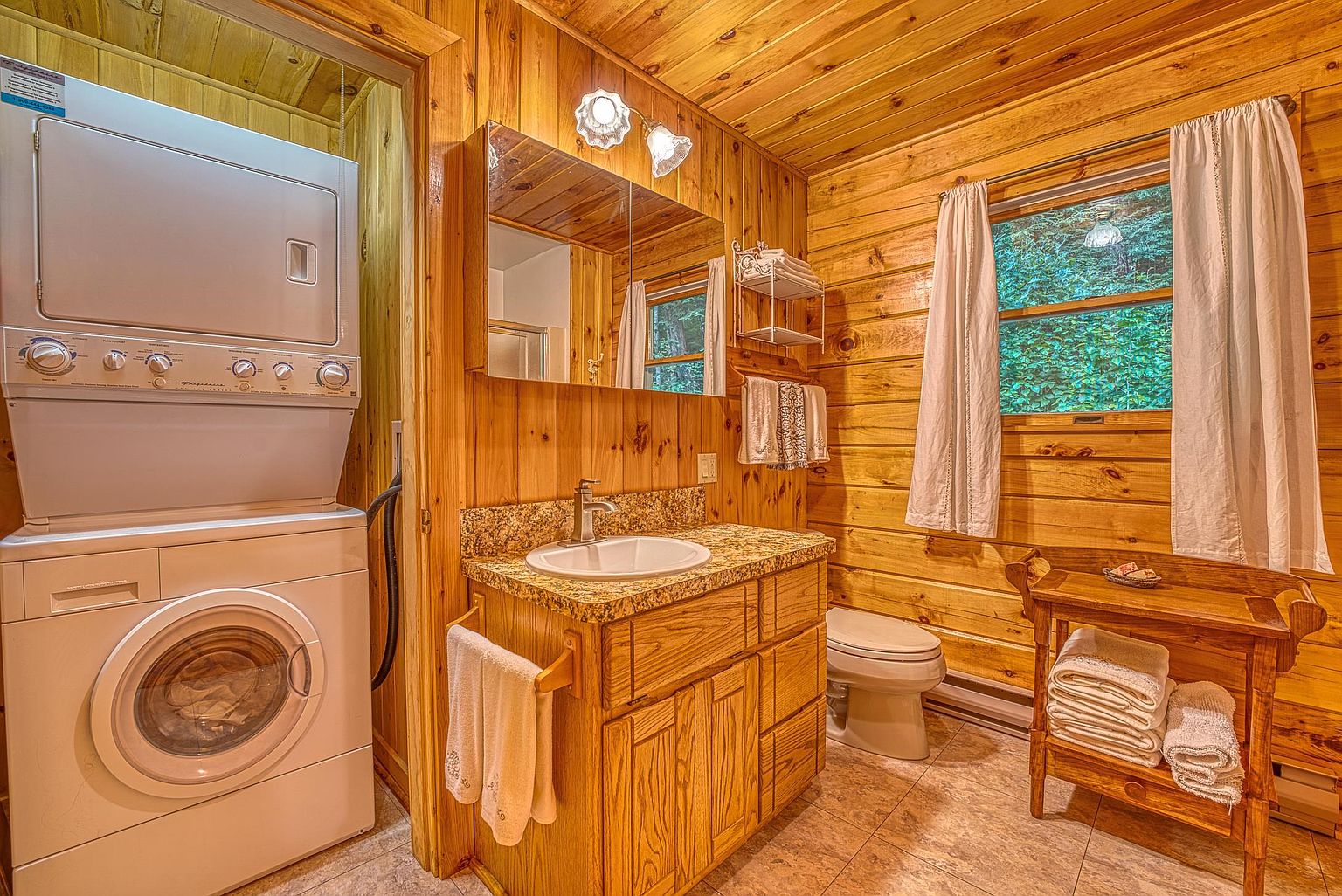 This rustic-style bathroom features warm wood-paneled walls and a stacked laundry unit integrated into the space. A vanity with a stone countertop and a single sink sits beneath a mirror, while a wooden stand provides storage for towels near a window with white curtains. The room is finished with neutral-toned floor tiles, creating a cozy and functional atmosphere.