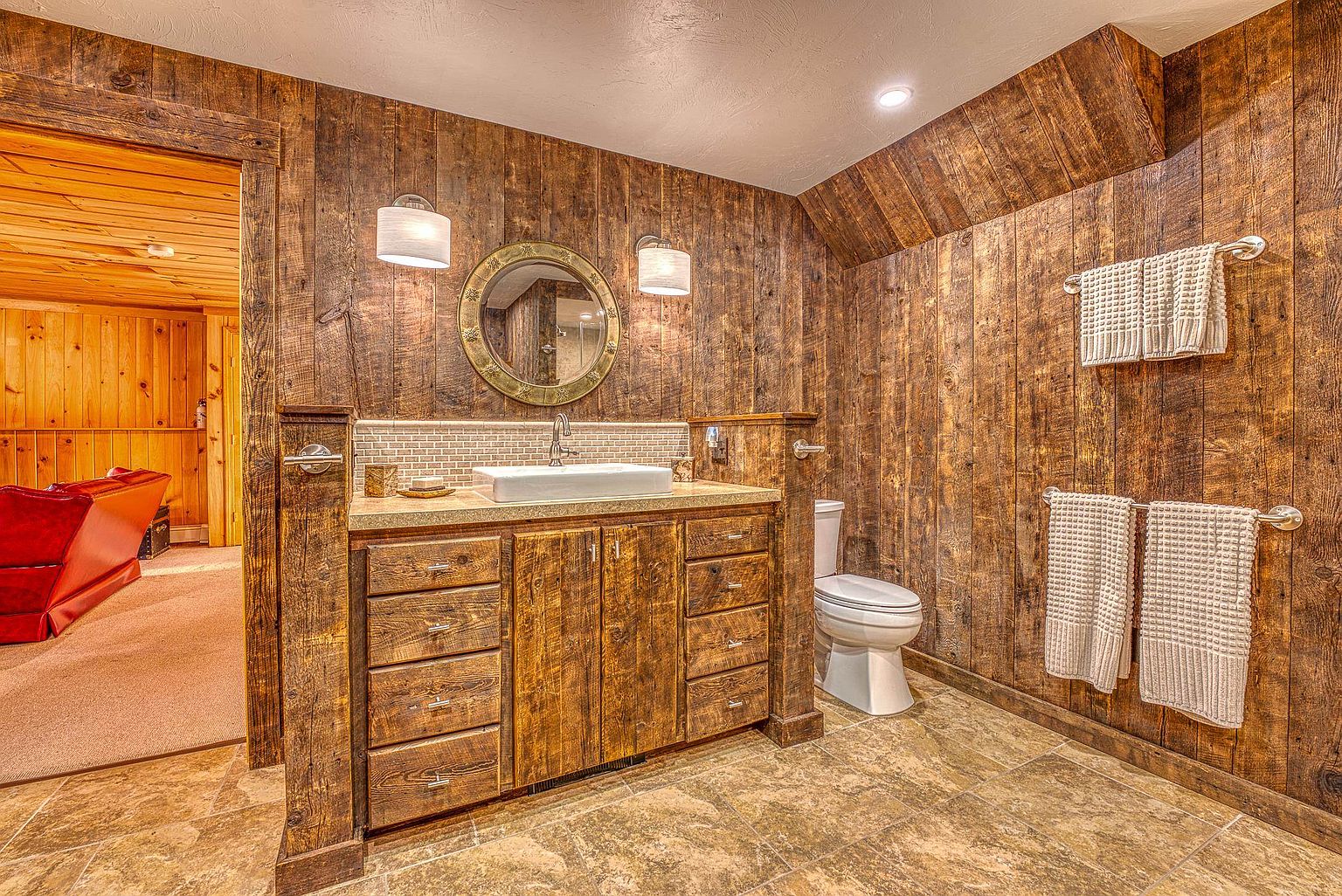 This rustic-style bathroom features warm wood-paneled walls and a matching custom vanity with a rectangular vessel sink. The space includes a toilet, a circular mirror with a decorative frame, and two wall-mounted light fixtures, all set against a backdrop of neutral-toned floor tiles. A doorway leads into an adjacent room, providing a glimpse of a red leather sofa and wood-paneled walls, enhancing the cozy, cabin-like aesthetic.