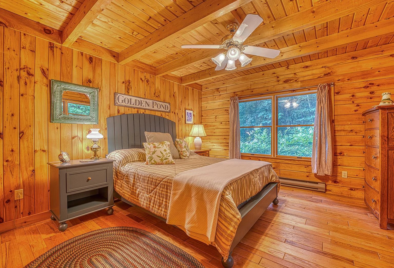 This cozy bedroom features warm, natural wood-paneled walls and a matching wood-beamed ceiling, creating a rustic cabin aesthetic. The room is centered around a bed with a dark-toned headboard, flanked by a matching nightstand and a tall wooden dresser. Large windows with light curtains allow natural light to filter in, while a ceiling fan and a braided rug add functional comfort to the space.