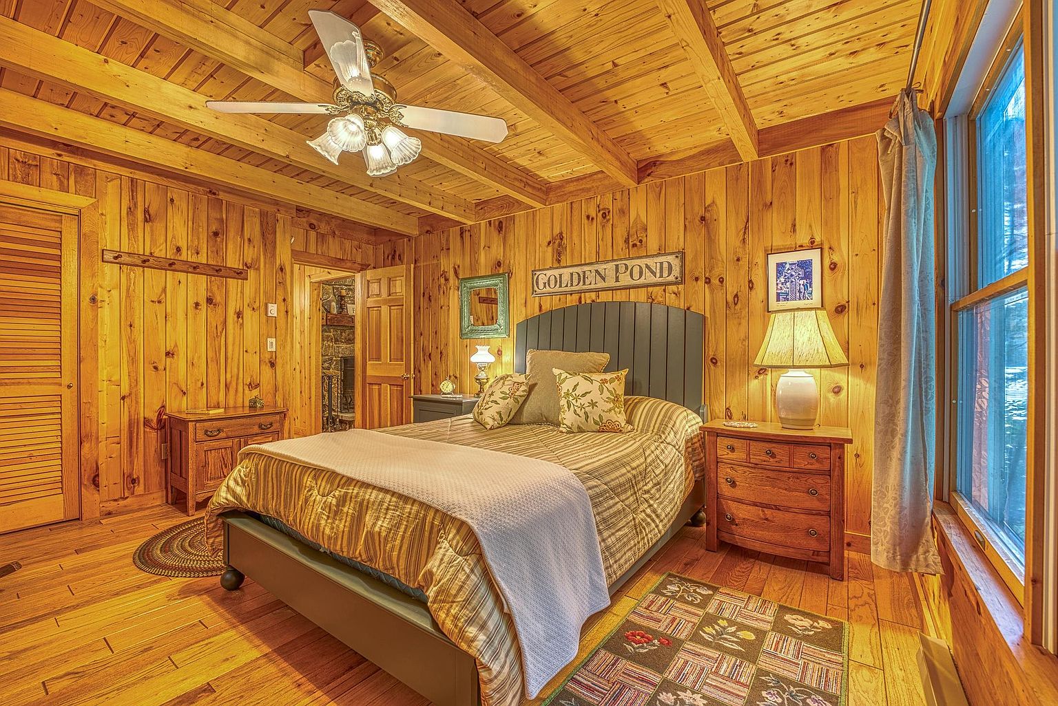 This cozy bedroom features warm wood-paneled walls and a matching wood-beamed ceiling, creating a rustic cabin atmosphere. A central bed with a dark headboard is dressed in plaid bedding, flanked by two wooden nightstands with lamps. The room is accented by a decorative 'Golden Pond' sign above the bed and a patterned rug on the wood floor, with a ceiling fan providing overhead lighting.