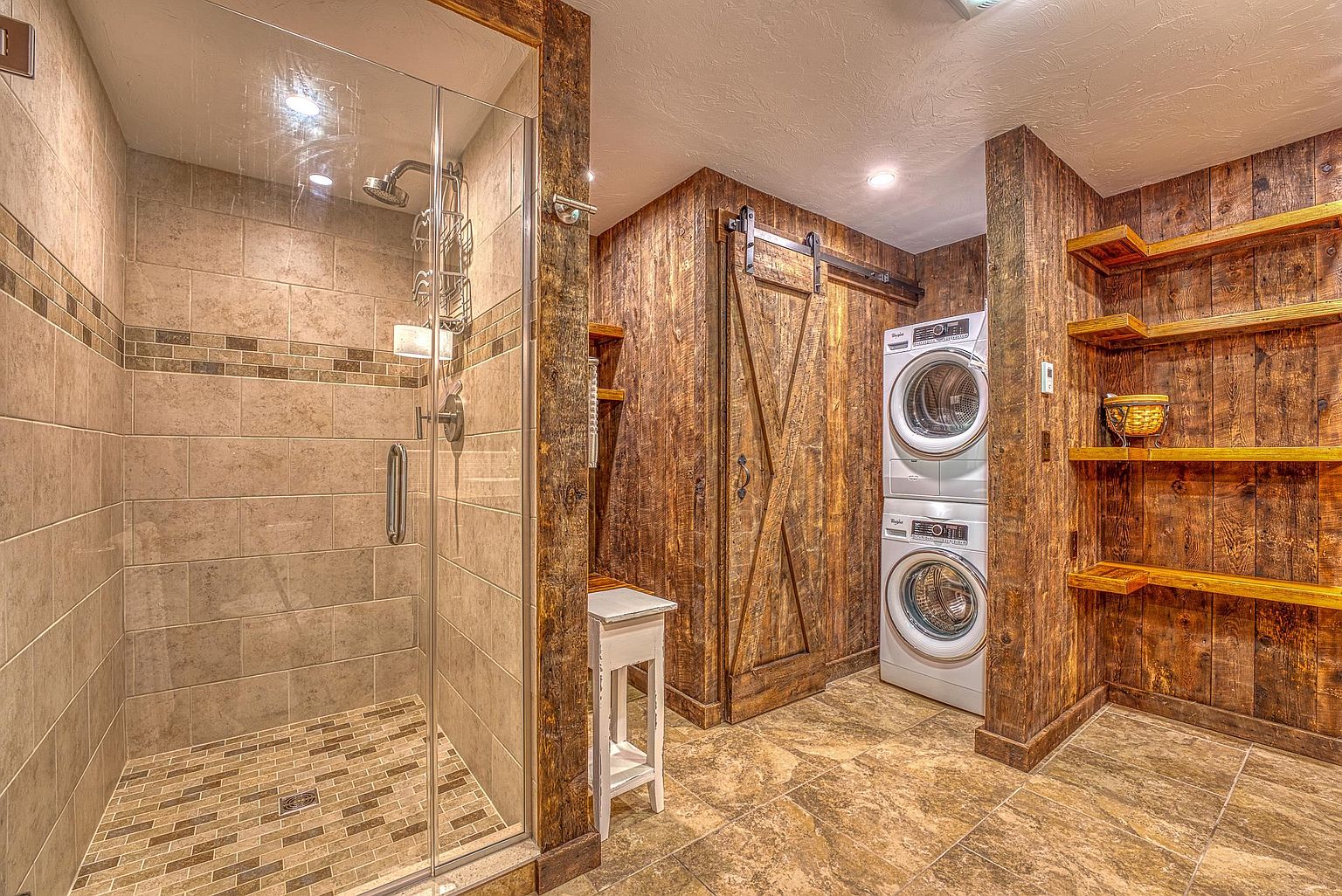 This rustic-style laundry room features a stacked washer and dryer unit set against a wall of reclaimed wood paneling. To the left, a glass-enclosed shower with neutral-toned tile adds a functional bathroom element to the space, while open wooden shelving on the right provides convenient storage. The room is finished with warm-toned floor tiles and a sliding barn-style door, creating a cohesive, cabin-inspired aesthetic.