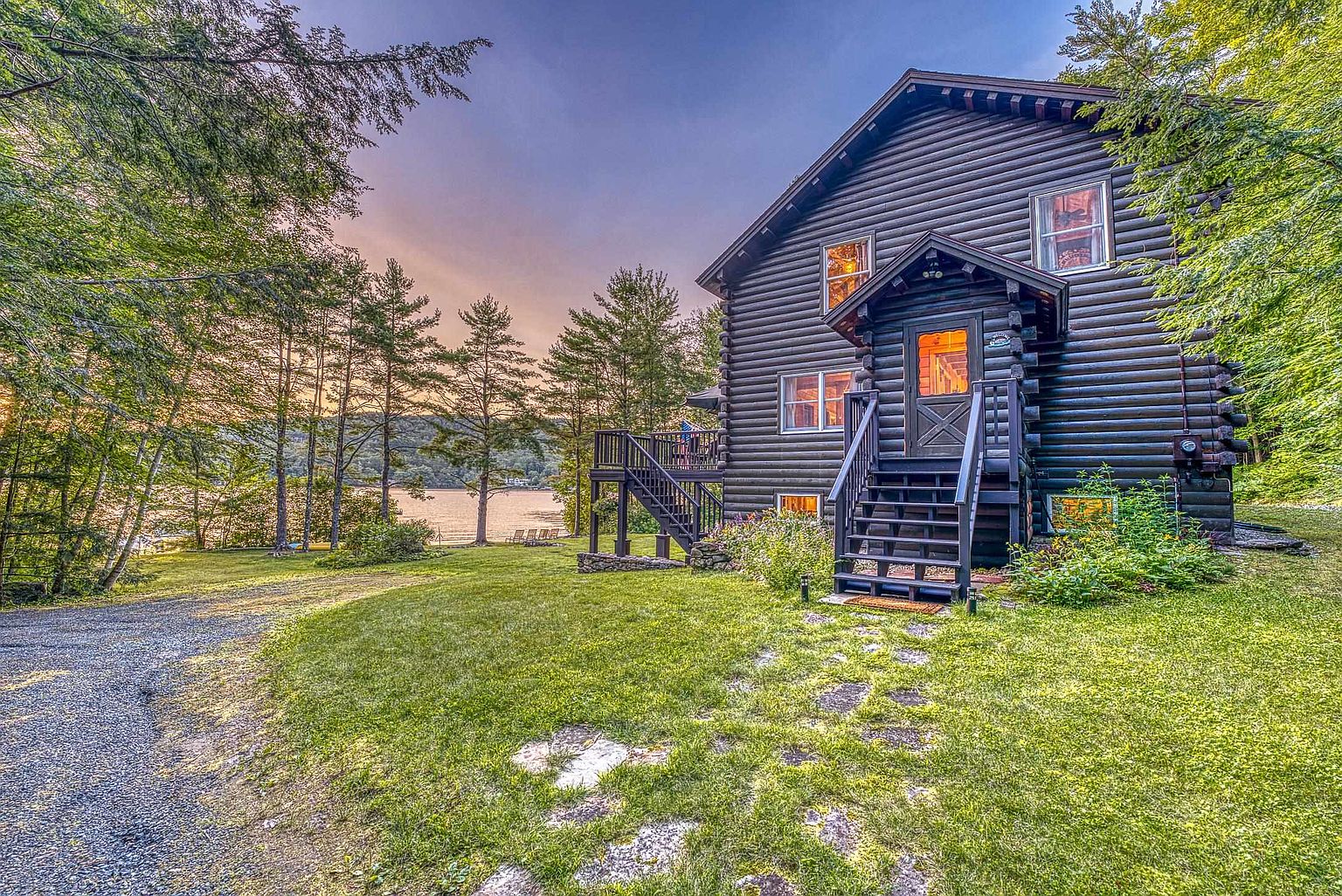 This charming log cabin features dark-stained horizontal timber walls and a prominent front entryway with a covered porch and wooden stairs. The home is nestled in a lush, green landscape with a gravel driveway leading toward a serene lake visible in the background under a soft, sunset-lit sky. The perspective captures the inviting, rustic aesthetic of the property, highlighting its integration with the surrounding mature trees and natural environment.