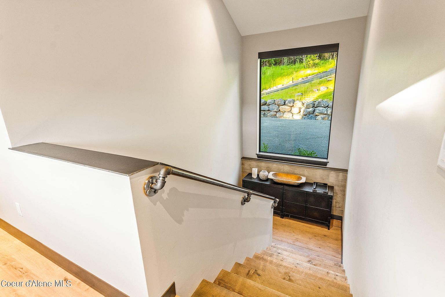 This interior shot showcases a modern staircase with wooden steps and a unique industrial-style metal handrail. A window at the landing provides natural light and a view of the exterior landscape. The walls are painted in a neutral tone, contributing to a clean and contemporary aesthetic.
