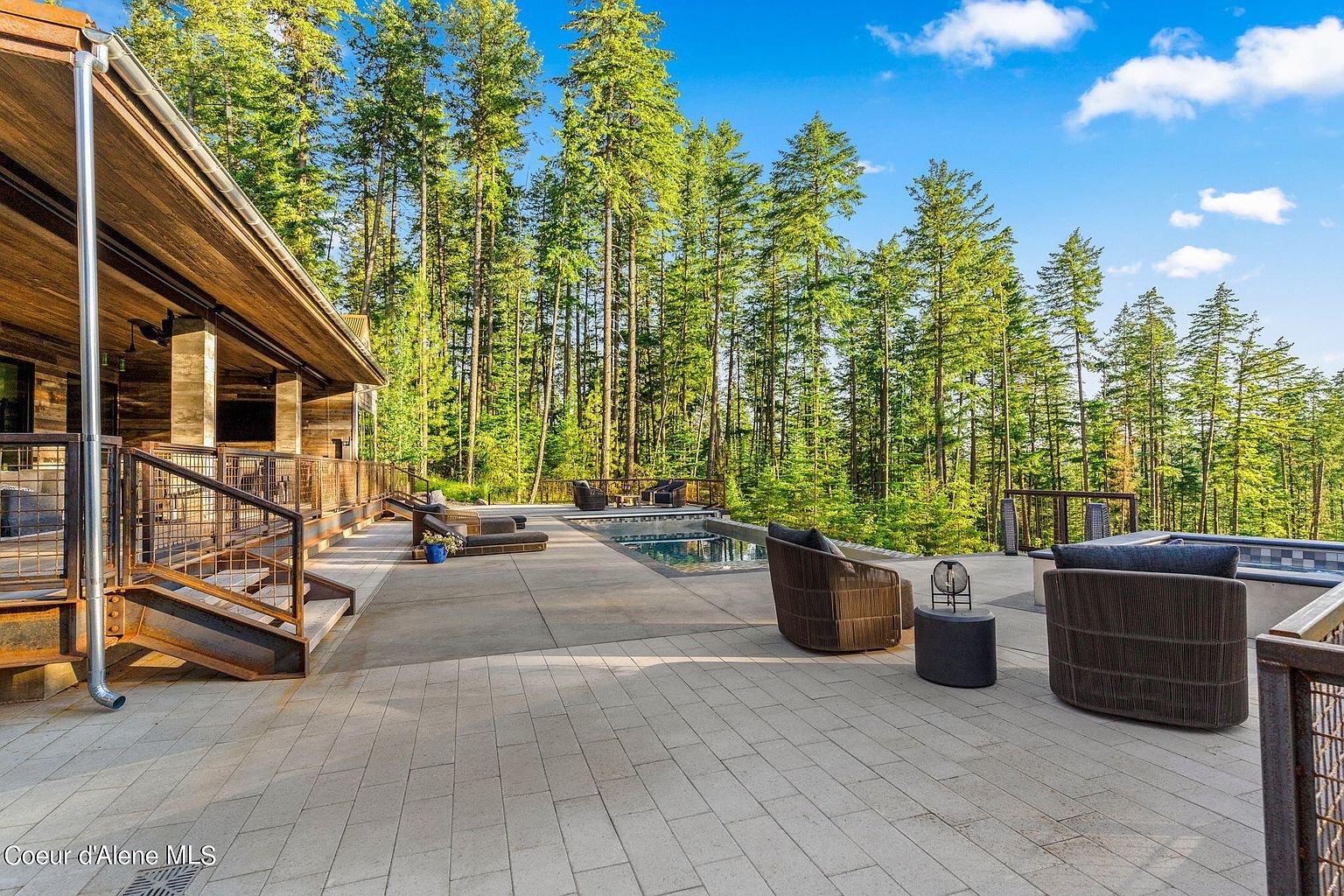 This image showcases a luxurious outdoor patio area, featuring comfortable wicker seating arrangements and a modern pool. The patio is constructed with light-colored pavers, and a wooden structure with metal railings adds architectural interest. Tall evergreen trees provide a scenic backdrop, creating a serene and private atmosphere.