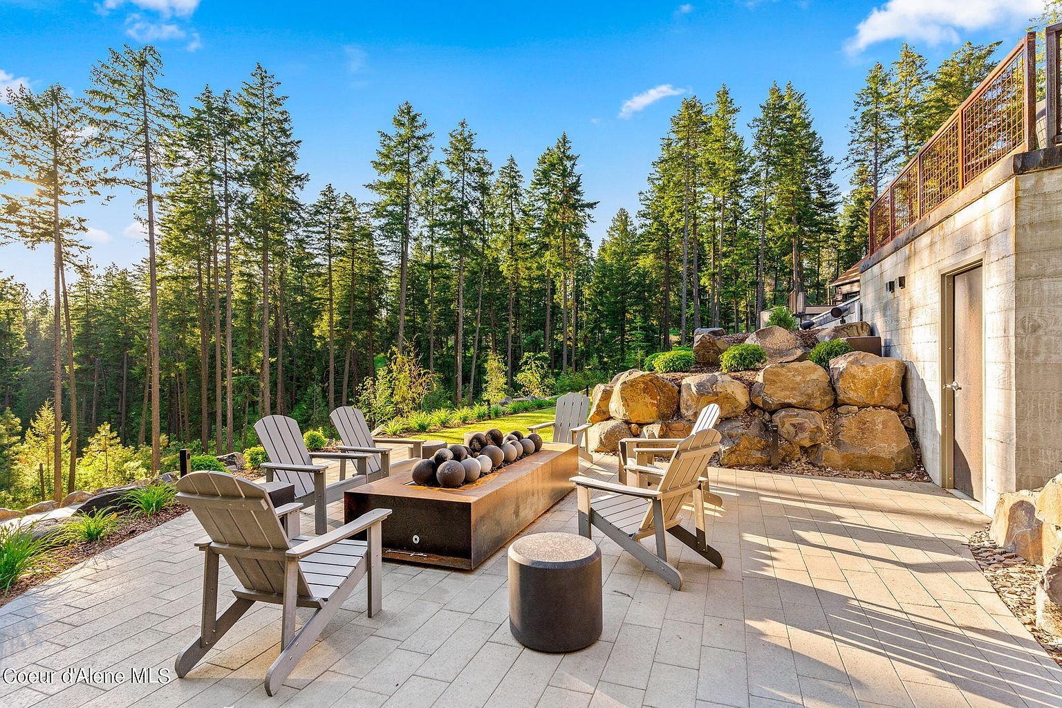 This image showcases a beautifully designed outdoor patio area, perfect for relaxation and entertainment. The space features a modern fire pit surrounded by comfortable seating, all set on a paved patio. The backdrop includes lush greenery and mature trees, creating a serene and private atmosphere. The patio is adjacent to a building with a stone foundation and a door, suggesting easy access to indoor spaces.