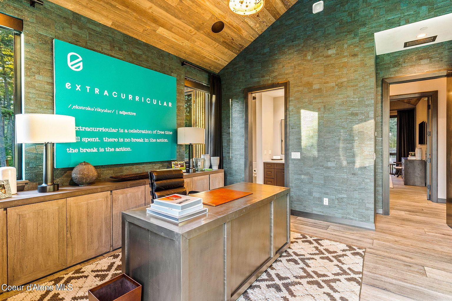 This is an interior shot of a modern office space featuring a large metal desk with an orange leather desk pad and stacked books. A teal wall sign with the word "EXTRACURRICULAR" adds a unique design element. The room is well-lit with a combination of natural light from the window and artificial light from the lamps, and the overall style is contemporary and sophisticated.