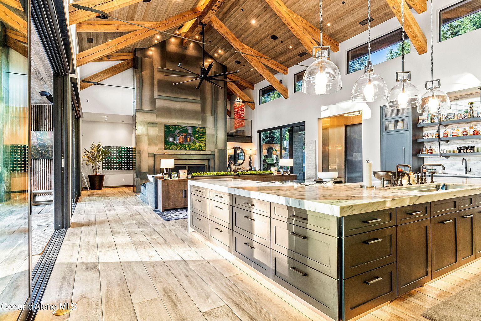 This is a spacious, modern kitchen with a large island featuring a marble countertop and dark cabinetry. The kitchen opens into a living area with a stone fireplace and high, exposed-beam ceilings. Natural light floods the space, highlighting the hardwood floors and creating a warm, inviting atmosphere.