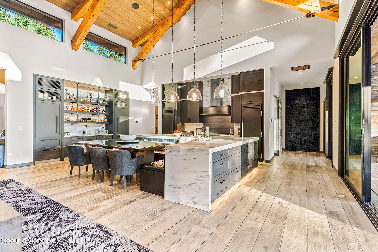 This is a spacious, modern kitchen featuring a large marble-topped island with seating, dark gray cabinetry, and stainless steel appliances. The kitchen also includes a dining area with gray upholstered chairs and a built-in bar area with open shelving. The room is well-lit with pendant lighting and natural light from clerestory windows, and the hardwood flooring adds warmth to the space.