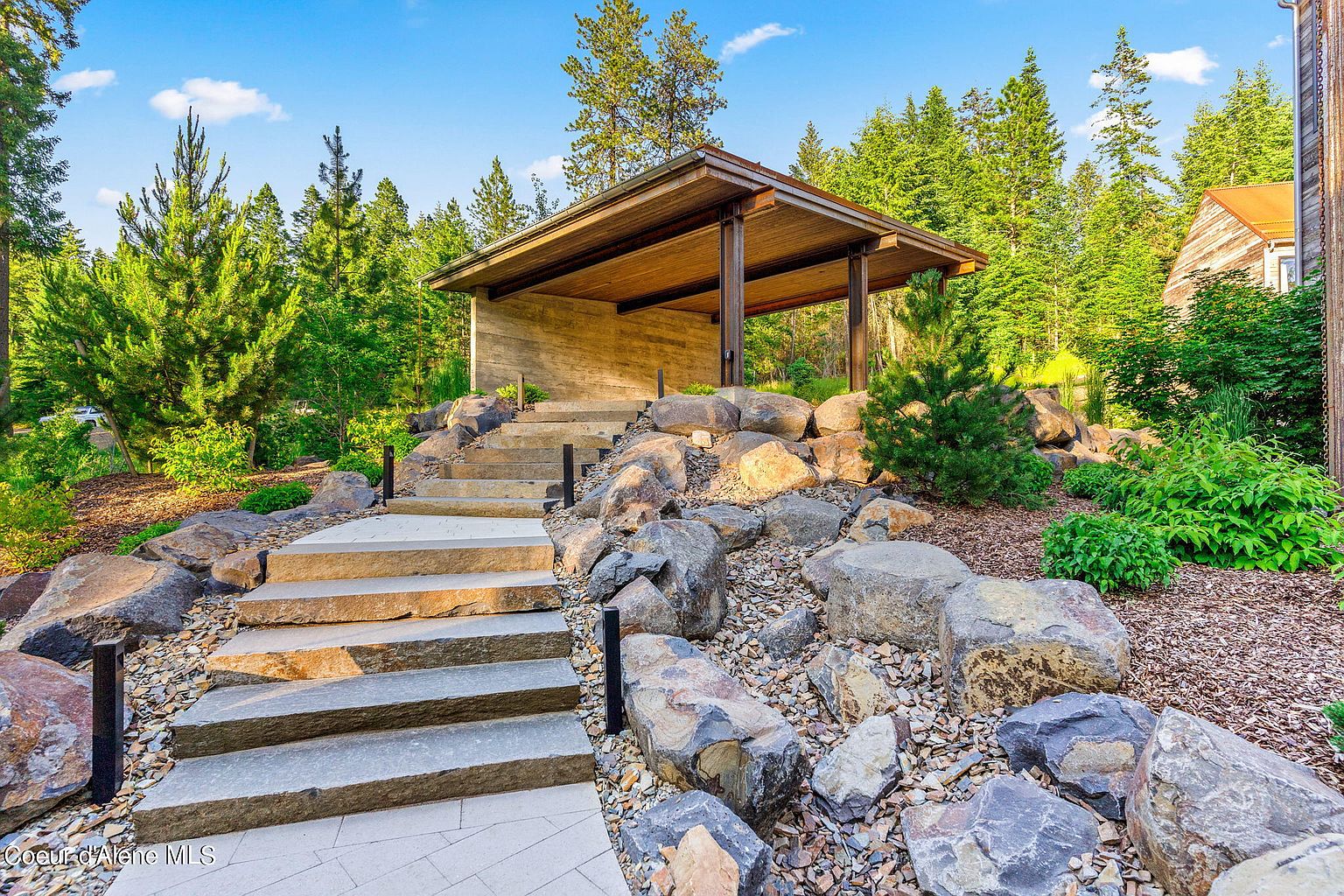 This image showcases a beautifully landscaped yard with stone steps leading up to a modern, open-air structure. The landscaping features a variety of rocks, plants, and trees, creating a natural and inviting atmosphere. The overall impression is one of tranquility and sophisticated outdoor living.