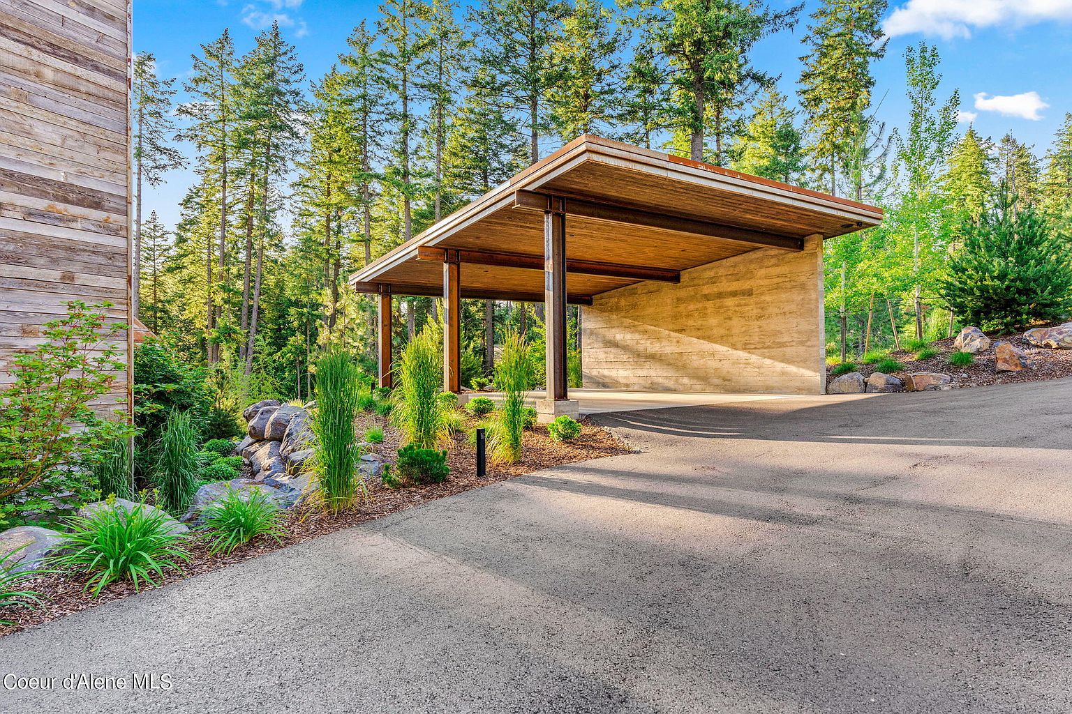 This image showcases a modern carport with a wooden roof supported by dark pillars, attached to a concrete wall. The driveway leads directly into the carport, surrounded by lush greenery and tall trees, creating a serene and private setting. The architectural style is contemporary and blends seamlessly with the natural landscape.