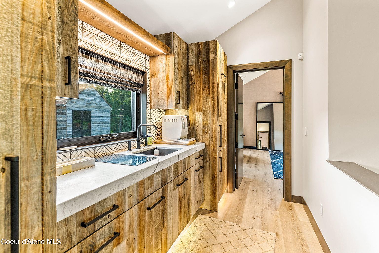 This interior shot showcases a rustic yet modern kitchen with wood-paneled walls and cabinetry, complemented by a light-colored countertop and black hardware. A window provides natural light and a view of the outdoors, while the open doorway leads to another room, creating a sense of flow. The overall design emphasizes natural materials and a warm, inviting atmosphere.