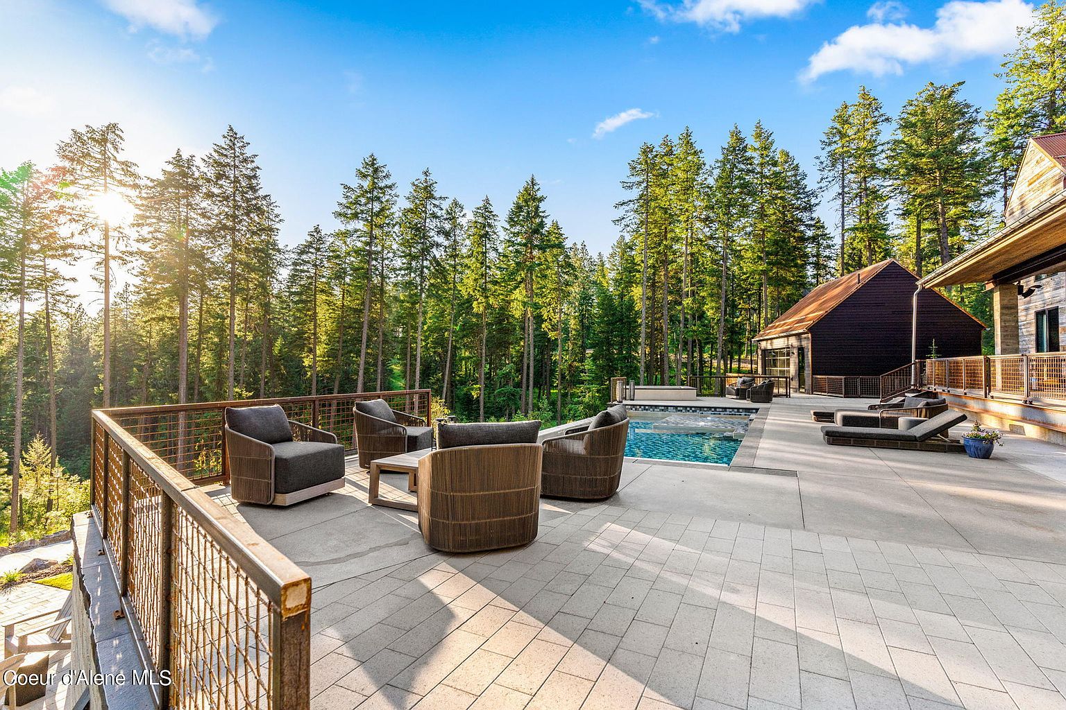 This image showcases a luxurious outdoor patio area, complete with comfortable seating arrangements, a swimming pool, and a backdrop of lush evergreen trees. The patio features modern furniture and a well-maintained deck, suggesting a perfect space for relaxation and entertainment. The adjacent building with its rustic wooden exterior adds to the property's charm and appeal.