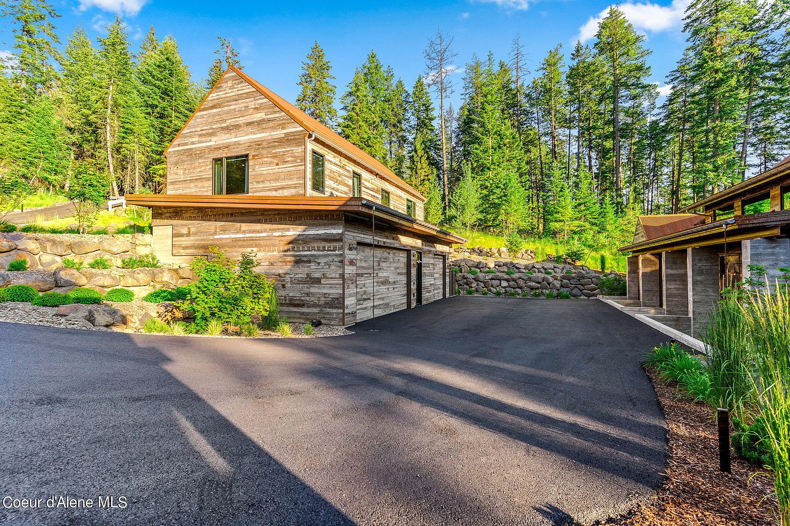 This image showcases the front exterior of a modern rustic home. The house features a combination of wood siding and stone accents, blending seamlessly with the surrounding wooded landscape. A paved driveway leads up to the house, and the overall impression is one of secluded luxury and natural beauty.