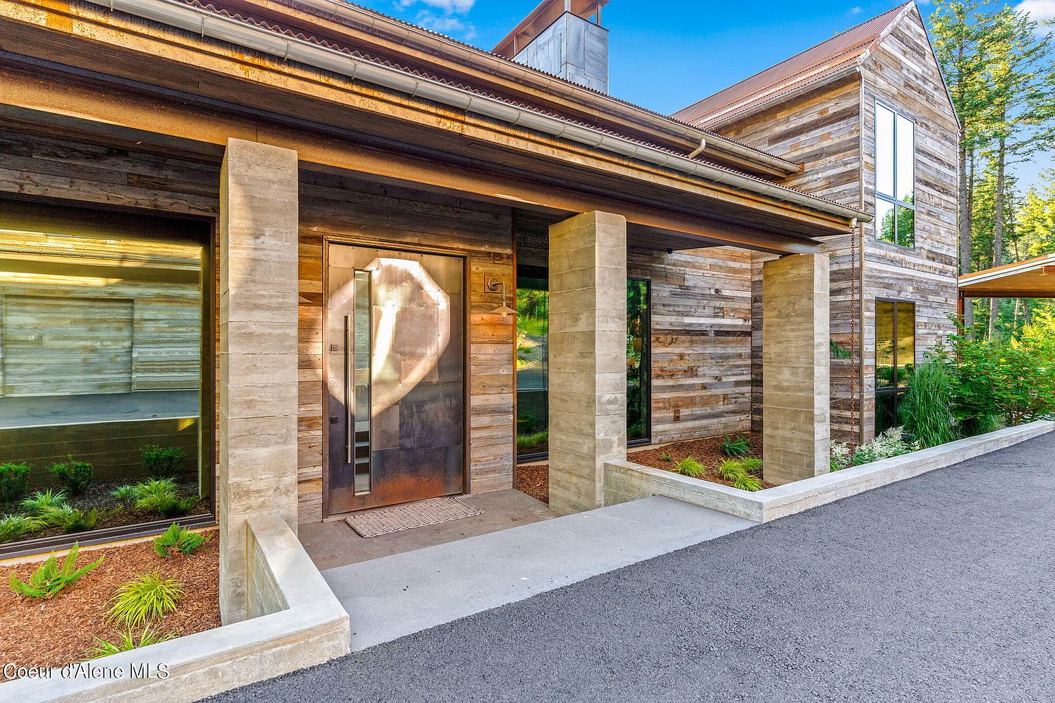 This image showcases the entryway of a modern home, featuring a unique metal front door with a striking design. The entrance is framed by concrete pillars and wood siding, creating a rustic yet contemporary aesthetic. A paved driveway leads up to the entrance, complemented by landscaped garden beds on either side.