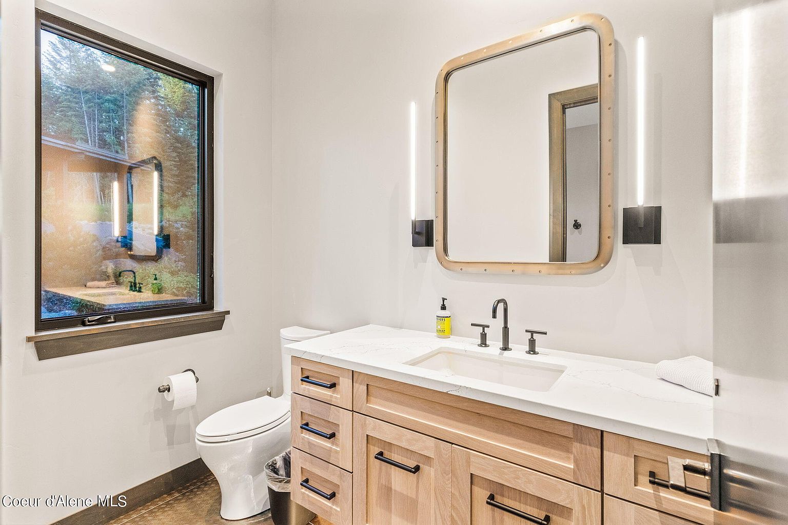 This is a well-lit guest bathroom featuring a modern design. The vanity has a light wood finish with black hardware and a white countertop with an integrated sink. A unique mirror with integrated lighting is mounted above the vanity, and a window provides natural light and a view of the outdoors.