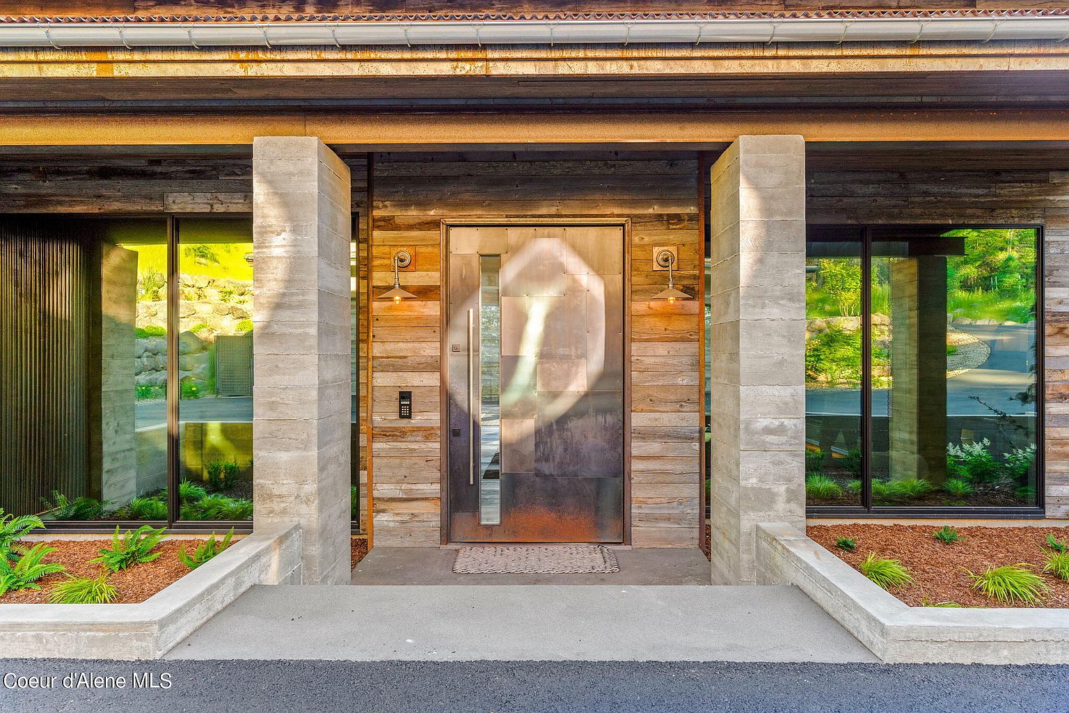 This image showcases the entryway of a modern home, featuring a striking metal front door flanked by concrete pillars and large windows. The door is set against a backdrop of horizontal wooden planks, with stylish sconces providing illumination. The landscaping includes neatly arranged plants and mulch, adding to the home's curb appeal.