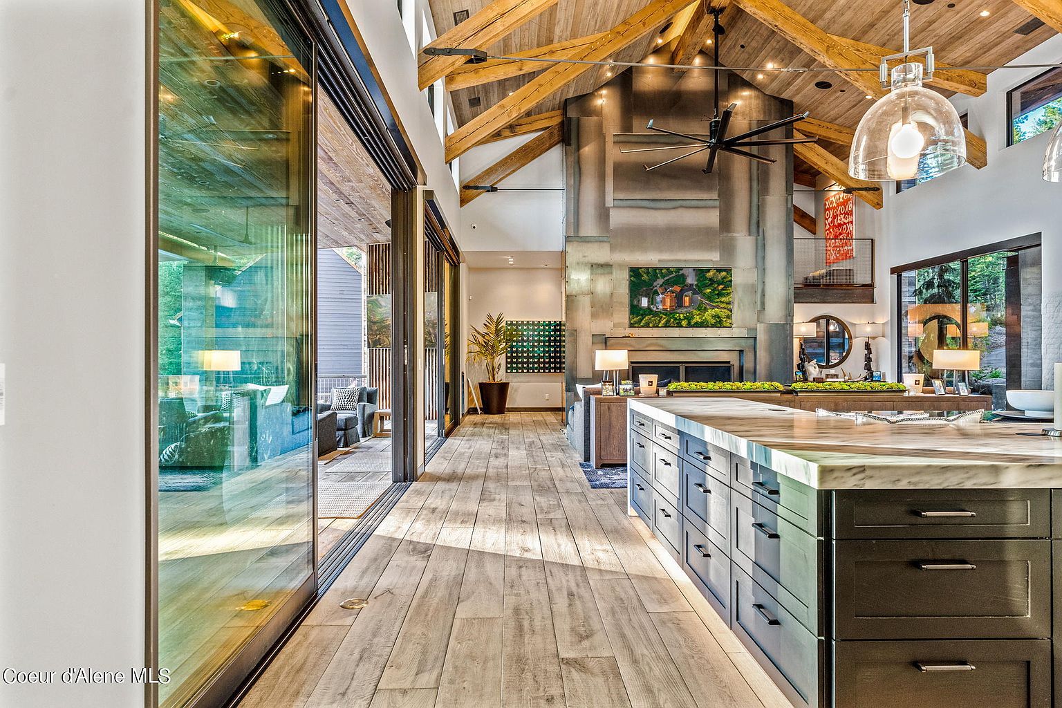 This interior shot showcases a spacious living area with a high, vaulted ceiling featuring exposed wooden beams. A large stone fireplace anchors the room, complemented by modern artwork and stylish furnishings. Expansive glass doors provide ample natural light and a seamless transition to an outdoor space, enhancing the home's open and airy feel.