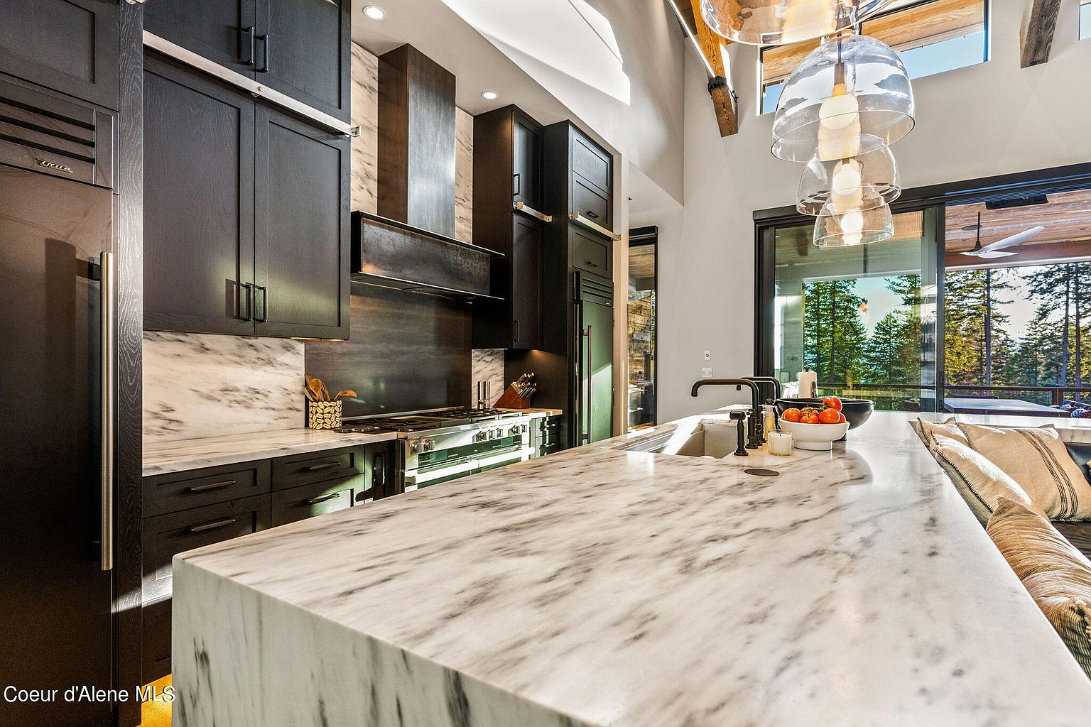This is a view of a modern kitchen with dark cabinetry and marble countertops. The kitchen features high-end appliances and a large island with a sink. Large windows provide natural light and a view of the outdoors, enhancing the space's appeal.