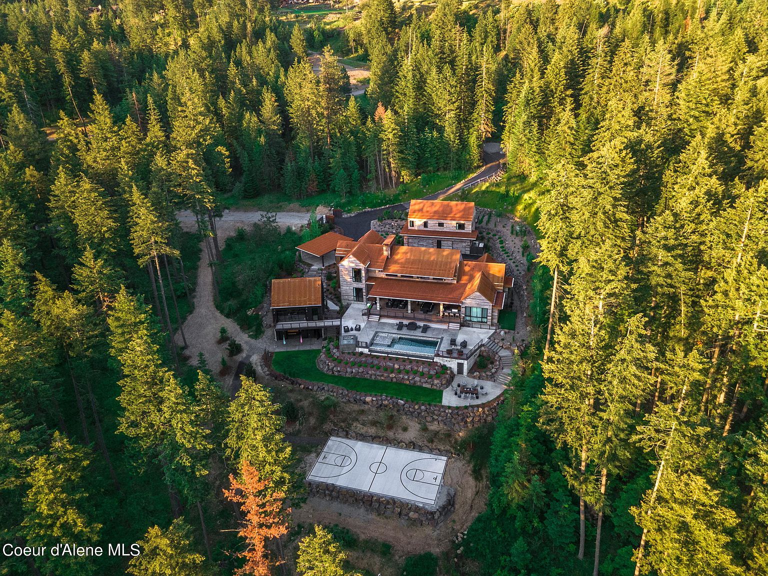 This aerial view showcases a luxurious estate nestled within a dense forest. The property features a multi-level home with a rust-colored roof, a swimming pool, a deck, and a basketball court. The surrounding landscape is lush with mature trees, providing privacy and a sense of seclusion.