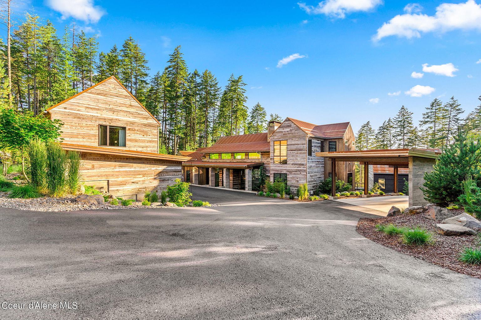 This is a front view of a modern rustic home with a unique architectural design. The exterior features a combination of wood siding and a metal roof, complemented by lush landscaping. A paved driveway leads up to the house, enhancing its curb appeal and highlighting the property's secluded, natural setting.