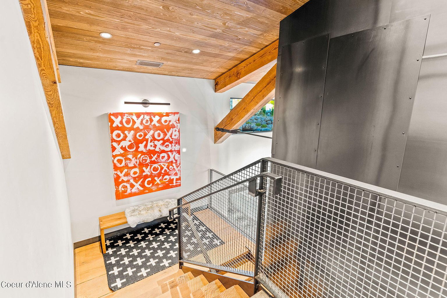 This interior shot showcases a modern hallway and staircase. The staircase features wooden steps and a metal grid railing, leading upwards. An orange and white abstract painting hangs on the wall above a small bench, adding a pop of color to the space. The ceiling is wood paneled with exposed beams, contributing to the contemporary design.