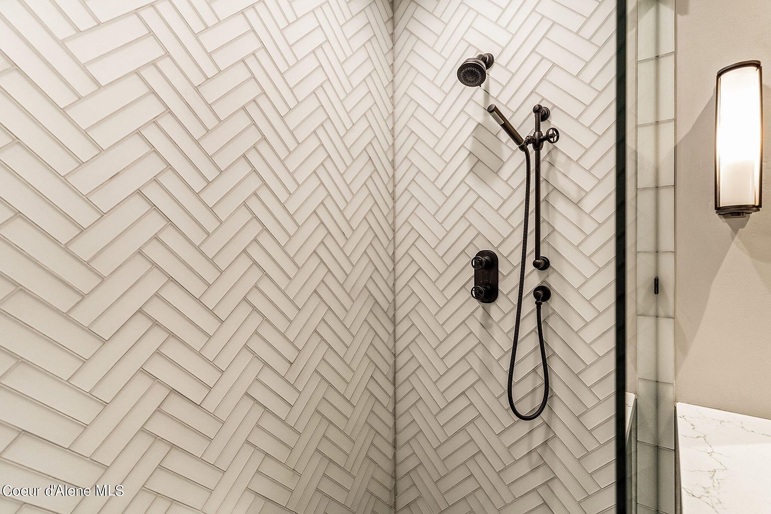 This is a detailed shot of a bathroom shower featuring a herringbone tile pattern on the walls. The shower is equipped with dark bronze fixtures, including a shower head and controls. A glass shower door is partially visible on the right, along with a sconce light fixture, adding a touch of elegance to the space.