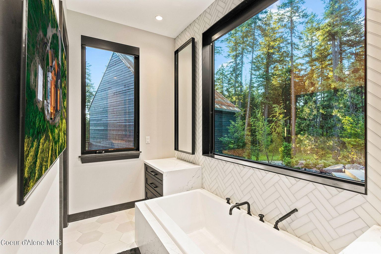This is a luxurious primary bathroom featuring a modern freestanding bathtub positioned beneath a large window offering a view of the surrounding trees. The bathroom is decorated with white herringbone tile and features black fixtures, adding a touch of elegance. A smaller window and a framed mirror are also visible, enhancing the natural light and spacious feel of the room.