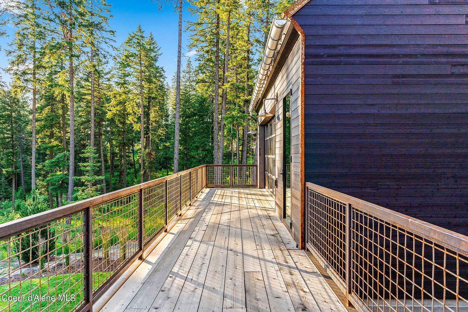 This image showcases a spacious wooden deck with metal railing, offering an inviting outdoor living space. The deck is adjacent to a modern home with dark wood siding and is surrounded by tall trees, creating a sense of privacy and connection with nature. The perspective is from the deck, looking out towards the trees.