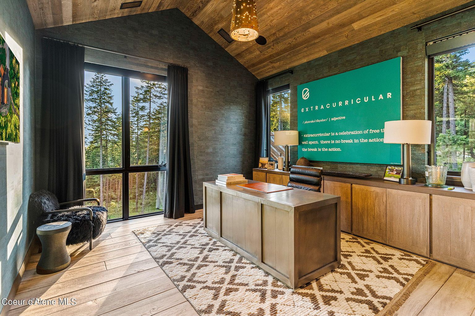 This is a well-appointed home office featuring a large desk, built-in cabinetry, and a statement wall with an 'extracurricular' graphic. The room has a modern rustic style with wood floors, a textured wall, and large windows offering a view of the surrounding trees. A comfortable chair and unique lighting fixture add to the room's appeal.