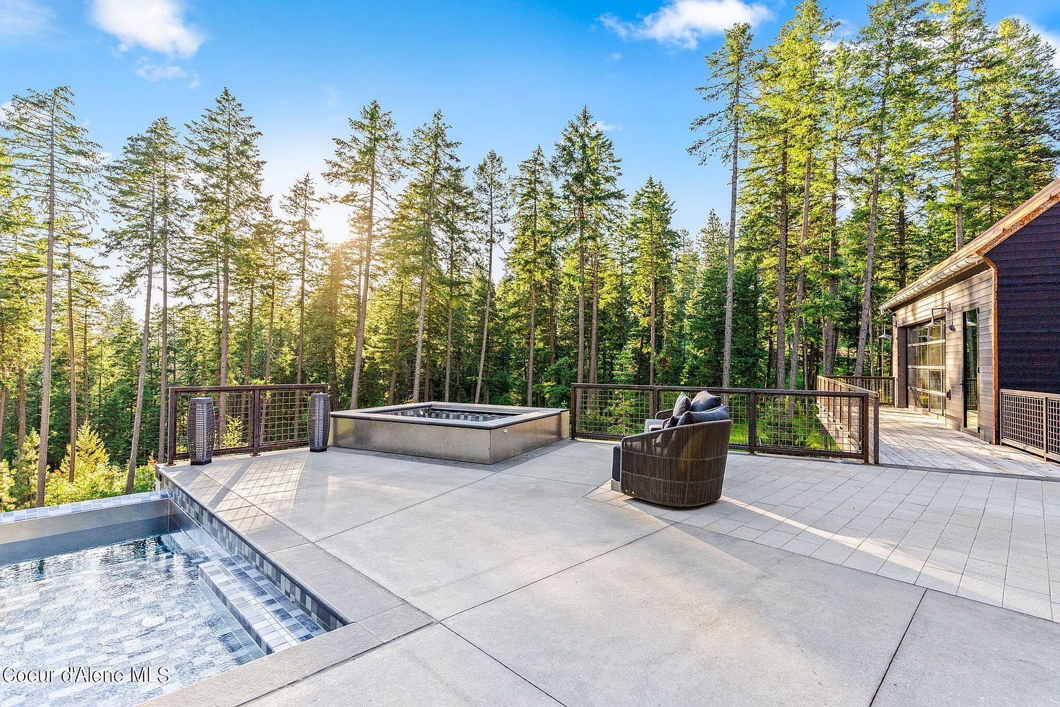 This image showcases a luxurious outdoor patio and deck area, complete with a hot tub, a comfortable lounge chair, and a swimming pool. The space is surrounded by tall trees, providing a sense of privacy and tranquility. The modern design of the deck and the natural setting create an inviting and upscale atmosphere, perfect for relaxation and entertainment.