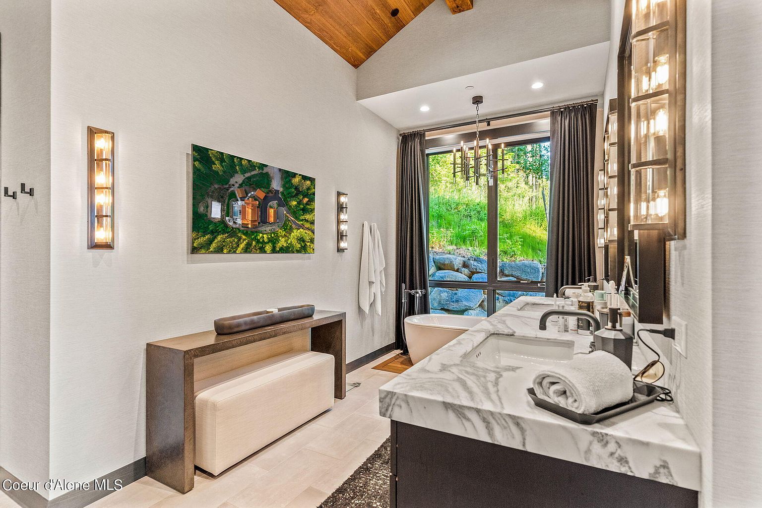 This is a luxurious primary bathroom featuring a double vanity with a marble countertop and dark wood cabinetry. A freestanding bathtub is positioned near a large window offering a view of the outdoors. The room is decorated with modern lighting fixtures and a console table with a bench, creating a spa-like atmosphere.