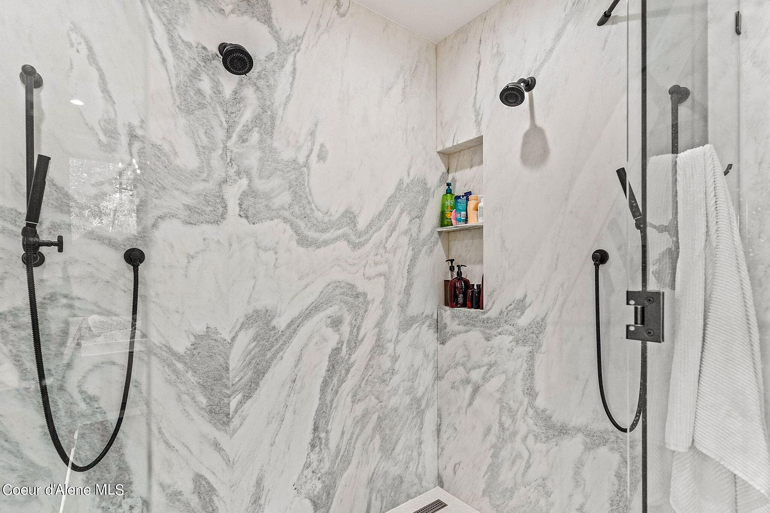 This is a well-lit primary bathroom showcasing a luxurious shower area. The walls are covered in marble with gray veining, creating a sophisticated and elegant look. The shower features dual shower heads and a built-in niche for toiletries, enhancing both functionality and aesthetic appeal.