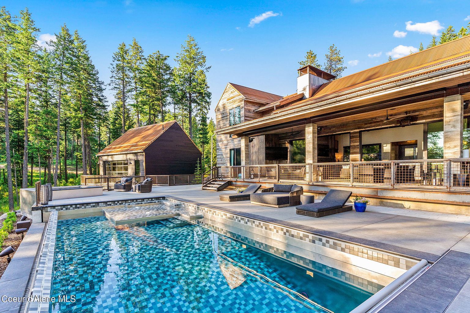 This exterior shot showcases a luxurious pool and spa area, seamlessly integrated with a modern rustic home. The pool features a mosaic tile design, and the surrounding patio includes lounge seating and a view of the wooded landscape. The home's architecture blends natural wood and metal accents, creating a sophisticated and inviting outdoor living space.