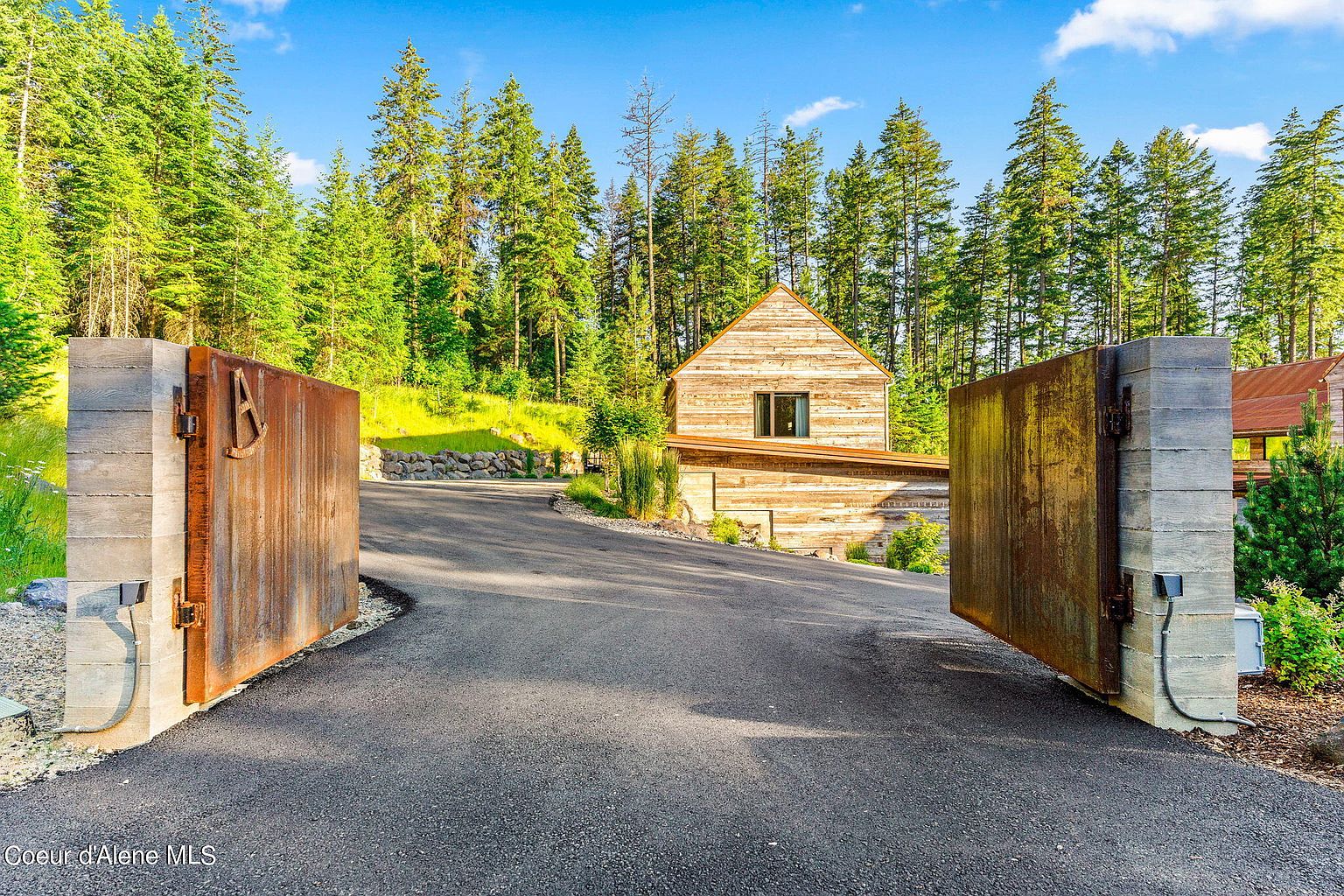 The image showcases a grand entryway to a property, featuring large, rustic metal gates flanked by concrete pillars. The driveway leads up to a wooden house nestled among tall evergreen trees under a bright blue sky. The scene evokes a sense of secluded luxury and natural beauty.