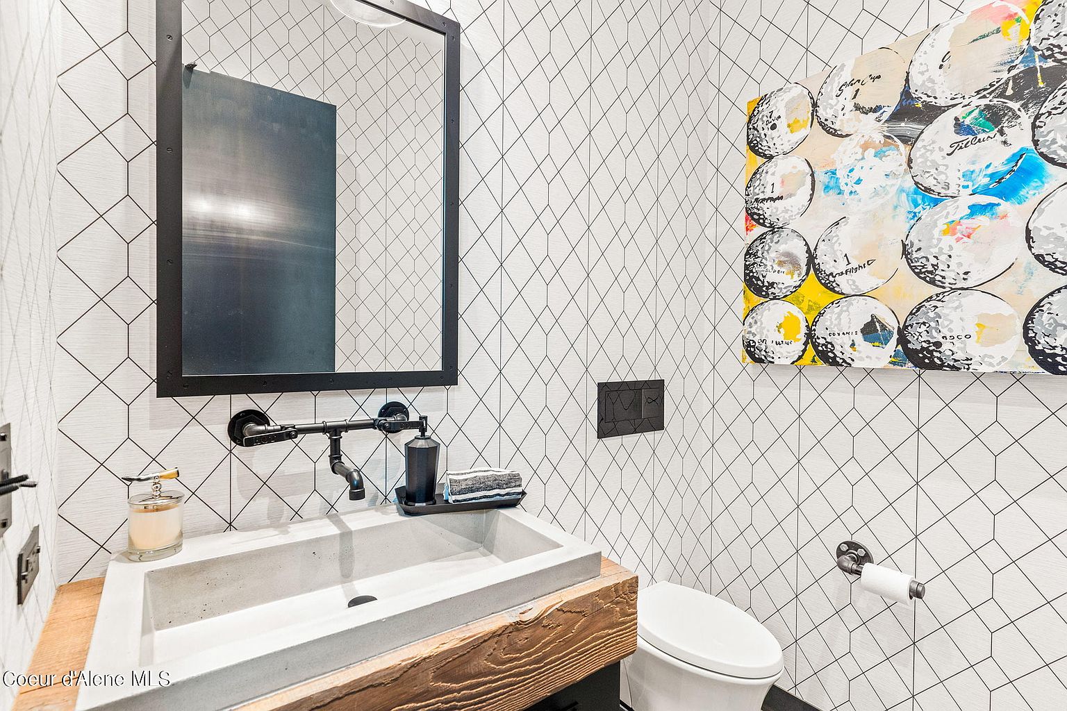 This is a modern guest bathroom featuring a unique geometric patterned wallpaper, a concrete sink with a wooden base, and black industrial-style fixtures. The artwork adds a pop of color, and the overall design is stylish and contemporary. The perspective is from the corner of the room, capturing the sink, toilet, and artwork.