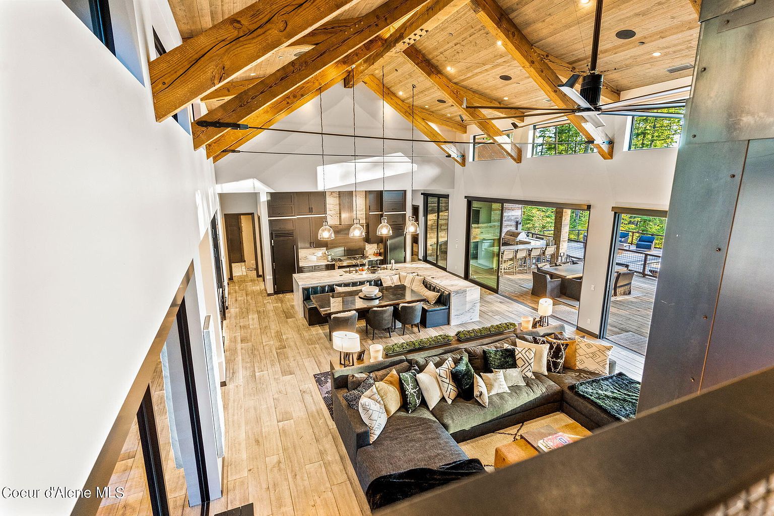 This is a high-angle shot of a luxurious living room and kitchen area, showcasing an open-concept design with exposed wooden beams and a soaring ceiling. The living room features a large sectional sofa adorned with decorative pillows, while the kitchen boasts a spacious island with seating and modern appliances. Sliding glass doors lead to an outdoor deck, seamlessly blending indoor and outdoor living spaces.