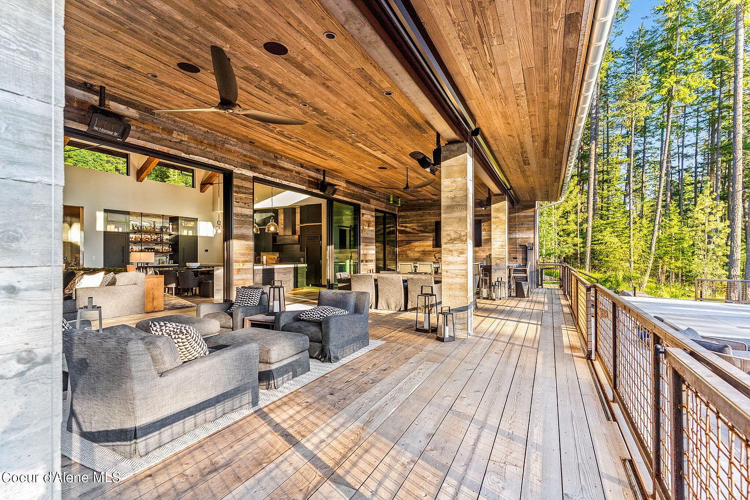 This image showcases a spacious outdoor patio/deck area, featuring comfortable gray lounge furniture arranged on a light-colored rug. The deck is constructed of wood and is partially covered by a wooden ceiling with a ceiling fan. The space is open to the interior through large sliding glass doors and offers views of the surrounding forest, creating a seamless indoor-outdoor living experience.