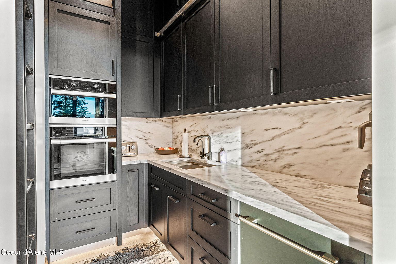 This is a modern kitchen featuring dark cabinetry and marble countertops and backsplash. The kitchen is equipped with stainless steel appliances, including a built-in oven and a dishwasher. The overall impression is sleek and sophisticated, with a focus on high-end finishes.