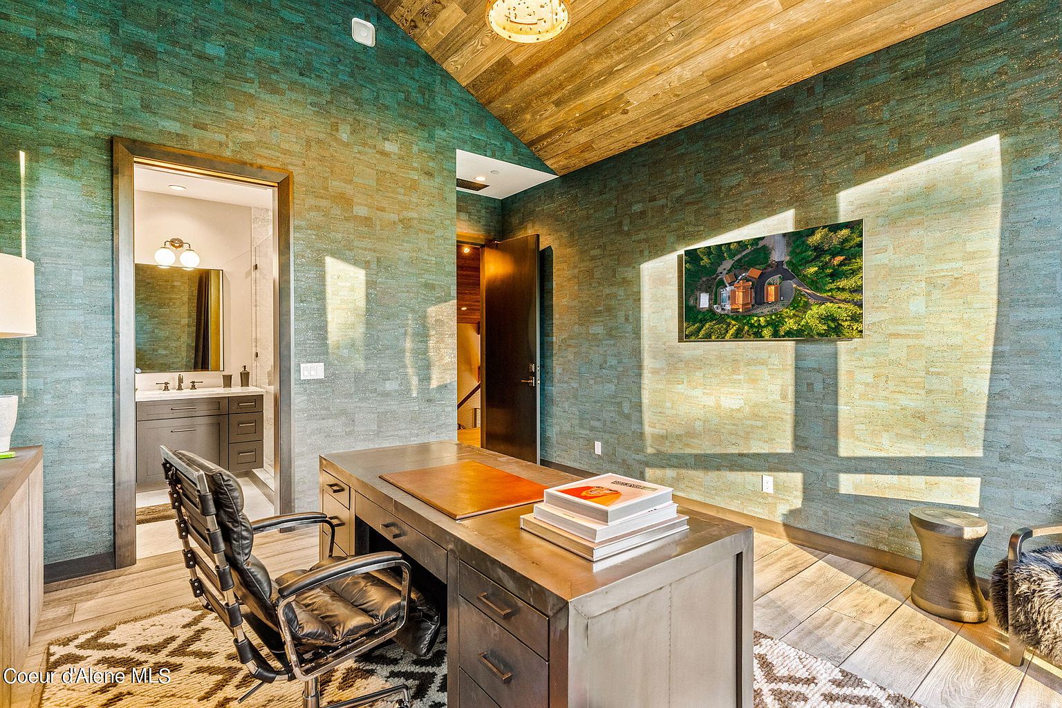 This is an interior shot of a modern office space featuring a unique textured wall, a wooden ceiling, and a large metal desk with books. A black leather chair sits in front of the desk, and a television displaying an aerial view of a property is mounted on the wall. The room has a sophisticated and stylish aesthetic, suitable for a professional or creative individual.
